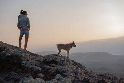 Woman with dog standing on rock against sky during sunset