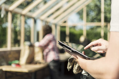 Cropped image of man using digital tablet while woman working in background at greenhouse