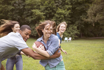 Man pulling woman holding football ball while friends running in background on field