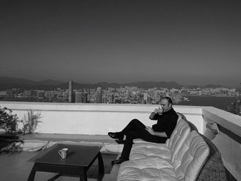 Man drinking coffee while sitting on sofa at building terrace against sky