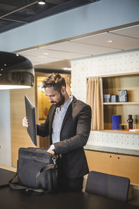 Young businessman keeping laptop in bag while standing by table in office