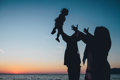 Silhouette parents playing with daughter standing by sea against sky at sunset