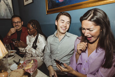 Happy women using smart phone while sitting by male friends at restaurant during party