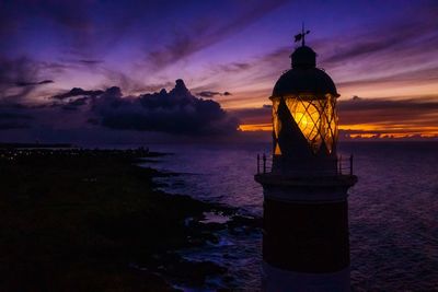 Illuminated lighthouse by sea against sky at sunset