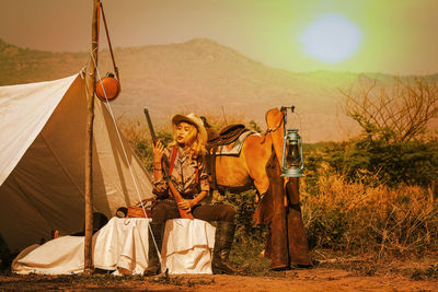 Woman sitting with gun outside tent with horse during sunset