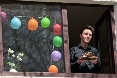 Low angle portrait of smiling teenage boy holding birthday cake at window