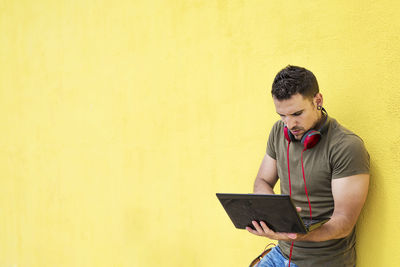 Side view of young man using laptop while standing against yellow background