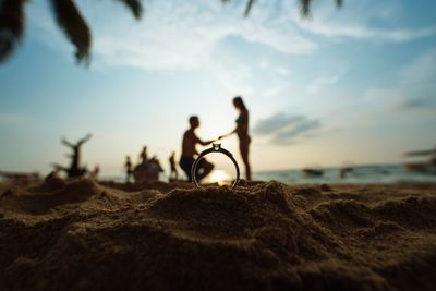 Close-up of ring in sand with couple in background at beach against sky