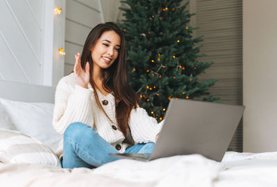 Young asian woman in white knitted sweater using laptop on bed in room with christmas 