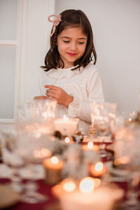 Cute girl in dress standing near festive table and lightning candles for celebrating christmas holiday