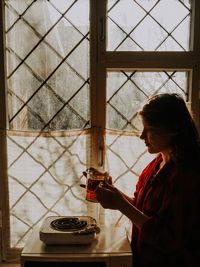 Side view of woman holding drink in glass by window at home