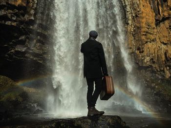 Rear view of man standing on rock while holding briefcase against waterfall