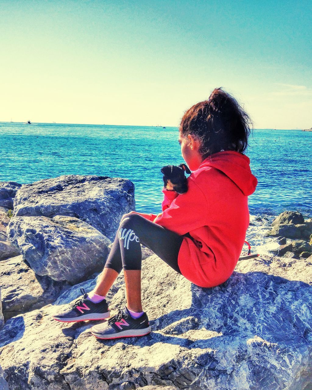 REAR VIEW OF WOMAN SITTING ON ROCK BY SEA