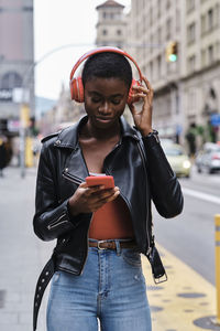 Woman listening music through headphones while using smart phone standing in city