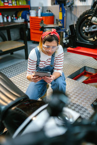 Concentrated female mechanic with security glasses holding digital tablet while review motorcycle