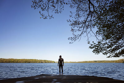 Rear view of female swimmer standing on boardwalk by lake against clear blue sky