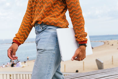 Side view of crop anonymous ethnic male in stylish colorful striped shirt and jeans carrying laptop while walking on embankment near sandy beach and sea