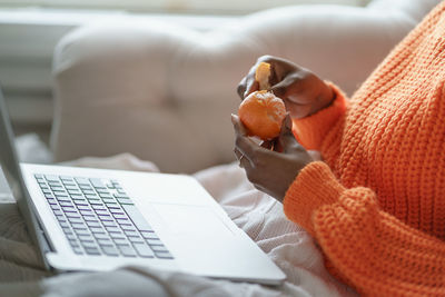 Hands peeling ripe tangerine, lying on couch under white knitted plaid, working on laptop at home. 
