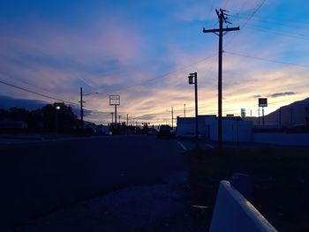 Road by silhouette electricity pylons against sky during sunset