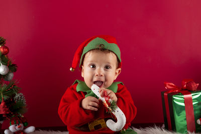 Sweet one-year-old boy dressed as an elf sitting, holding a typical christmas stick in his mouth