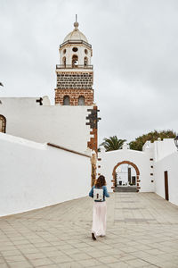 Back view of anonymous female with backpack walking on pavement against white houses and cloudy gray sky on town street in fuerteventura, spain