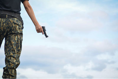 Midsection of soldier holding handgun against cloudy sky