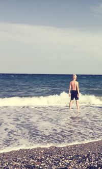 Rear view of shirtless teenage boy standing amidst surf on shore