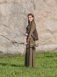 Portrait of woman holding bow and arrow standing on grass against rock