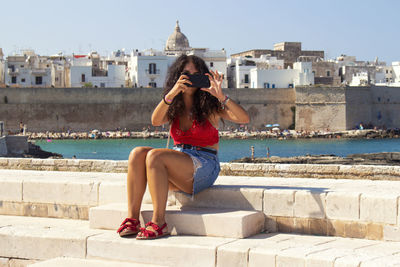 Woman photographing while sitting on retaining wall sea 