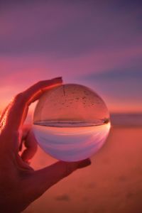 Close-up of hand holding crystal ball against sky during sunset