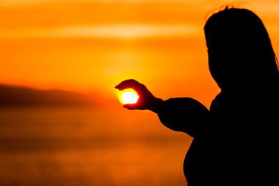 Optical illusion of silhouette woman holding sun at beach during sunset