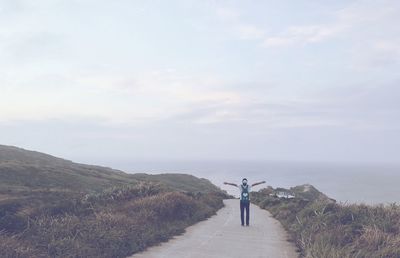 Rear view of man with arms outstretched standing on road against cloudy sky