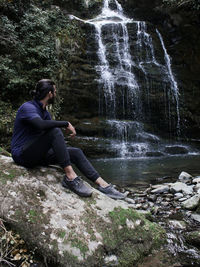A young adult sitting on a rock right next to a waterfall.