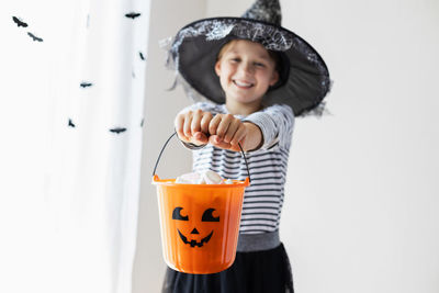 An orange pumpkin bucket full of sweets is being held by a charming girl dressed as a witch. 