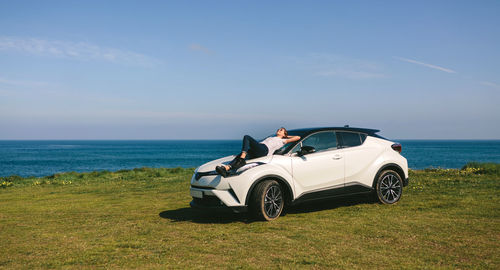 Full length of woman lying on car at field against blue sky