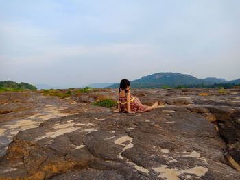 Rear view of girl sitting on rock against sky