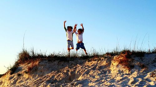 Low angle view of brothers jumping on sand hill at beach against clear sky