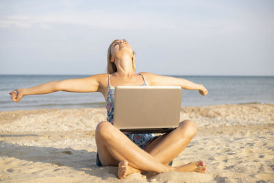 Young woman using laptop at beach against sky