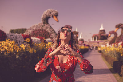 Young woman puckering lips while making heart shape standing in ornamental garden at dusk