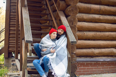 Man and a woman in red hats sitting on the stairs of wooden house in autumn and covered with a scarf