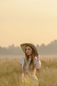 Portrait of woman wearing hat standing on field against sky during sunset