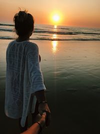 Woman holding cropped hand at beach against sky during sunset