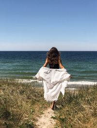 Rear view of woman undressing while walking at beach against blue sky