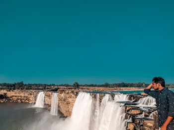 Young man standing by waterfall against clear sky