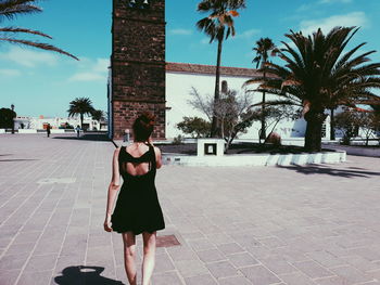 Woman standing on footpath by palm trees against sky