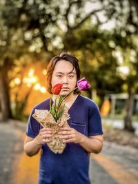 Portrait of young asian man holding red and purple tulips against trees and setting sun.