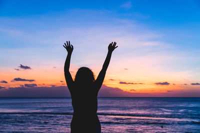 Silhouette woman with arms raised standing against sea during sunset