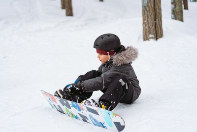 Little boy sitting on snow putting his feet in snowboard bindings adjusting straps