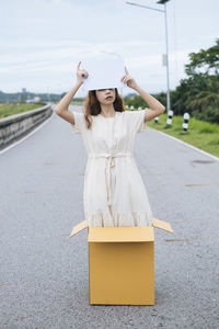 Young woman holding placard while standing in cardboard box on road