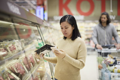 Woman doing shopping in supermarket and using cell phone to compare prices or checking shopping list
